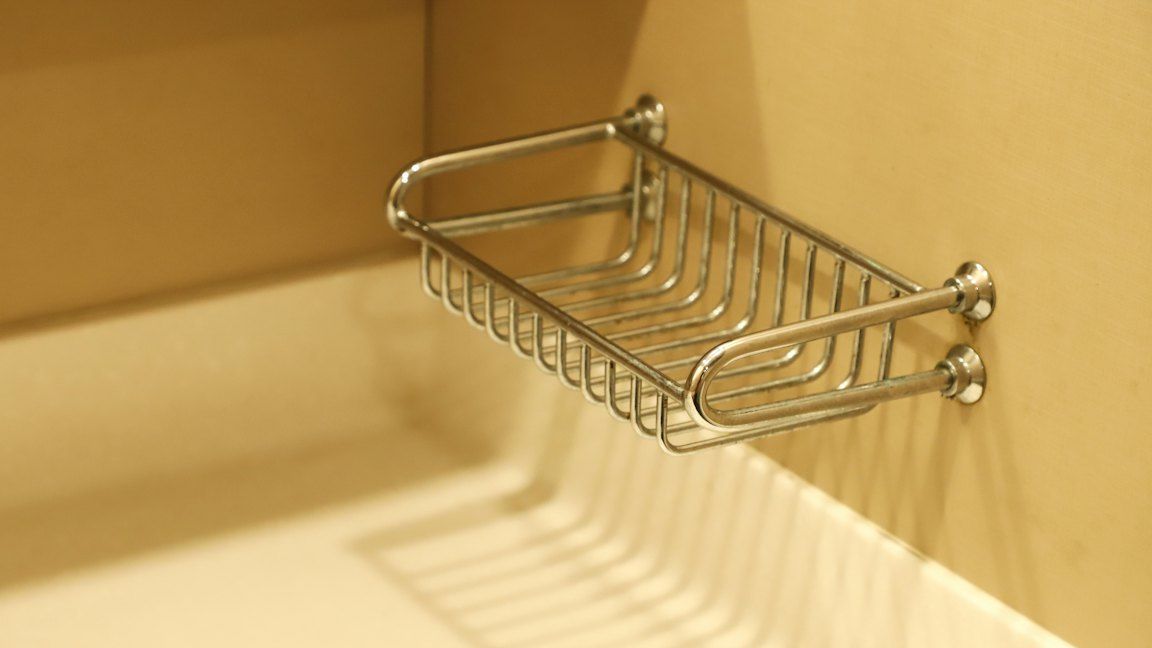 A chrome wire basket fixed to a beige wall, above the edge of a bathtub.