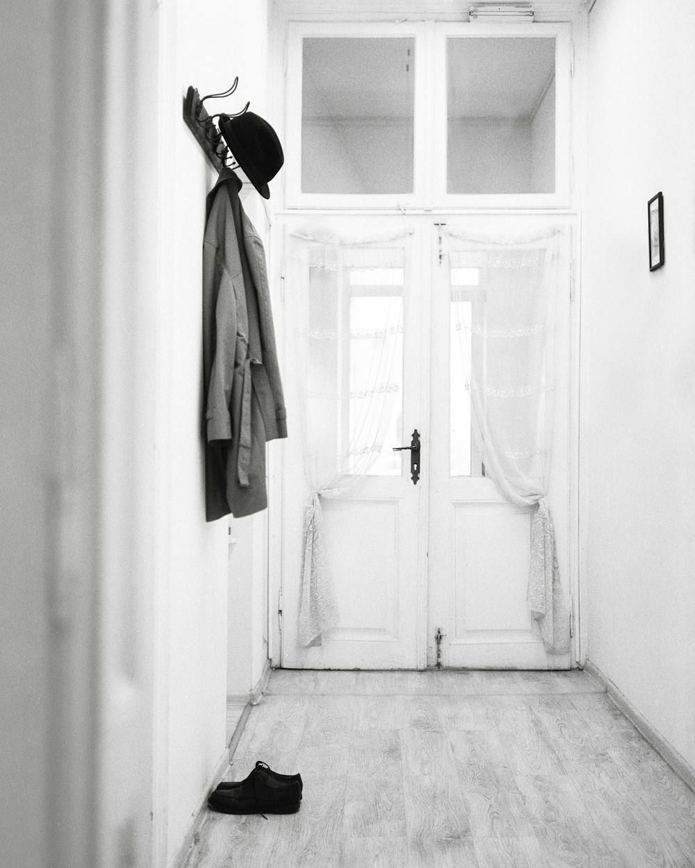 In a white hallway, in black and white, a coat and a bowler hat hang from a row of hooks on the left wall; a glazed door fills the far end and a pair of shoes stands on the floor.