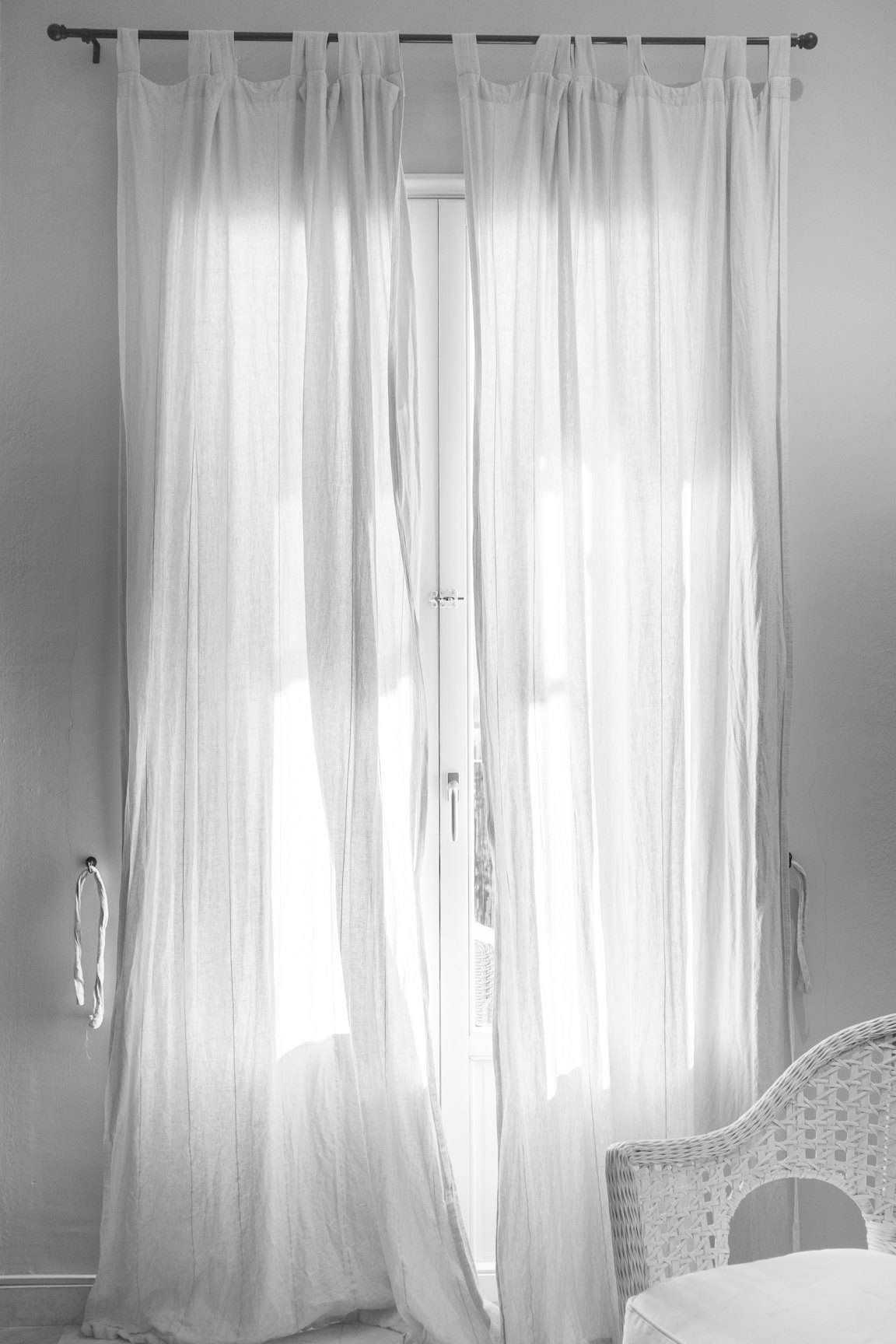 Two white tab-top curtains hanging from a thin black rod in front of a French window, in black and white.