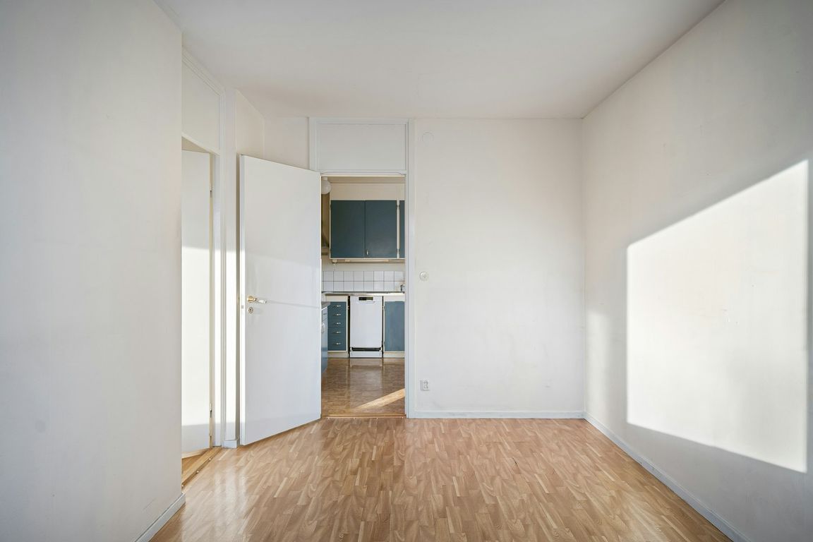 An empty room with white walls and a light wood floor, with an open door onto a kitchen and a patch of sunlight on the right-hand wall.