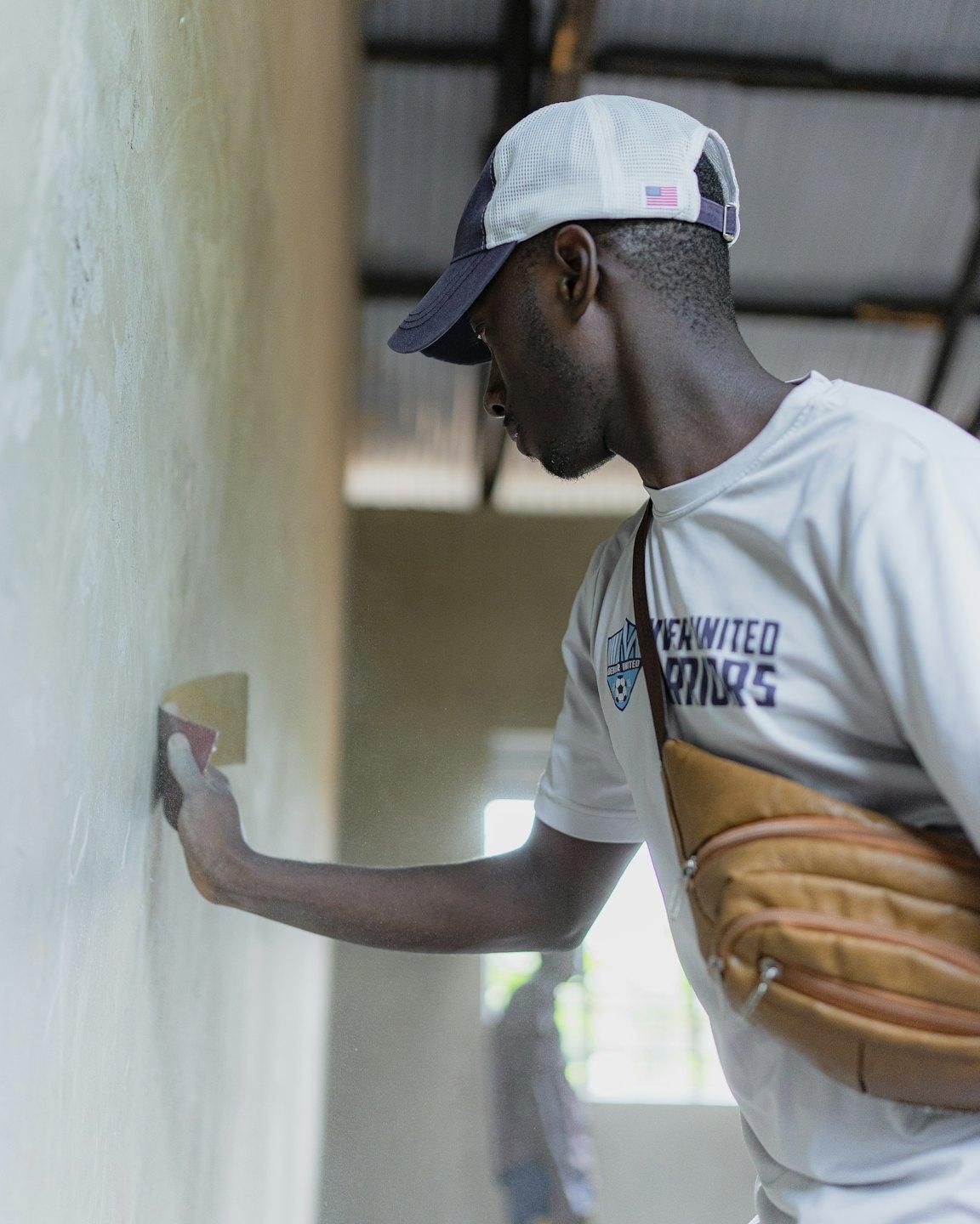 A man in a cap sands a wall by hand with a piece of sandpaper.