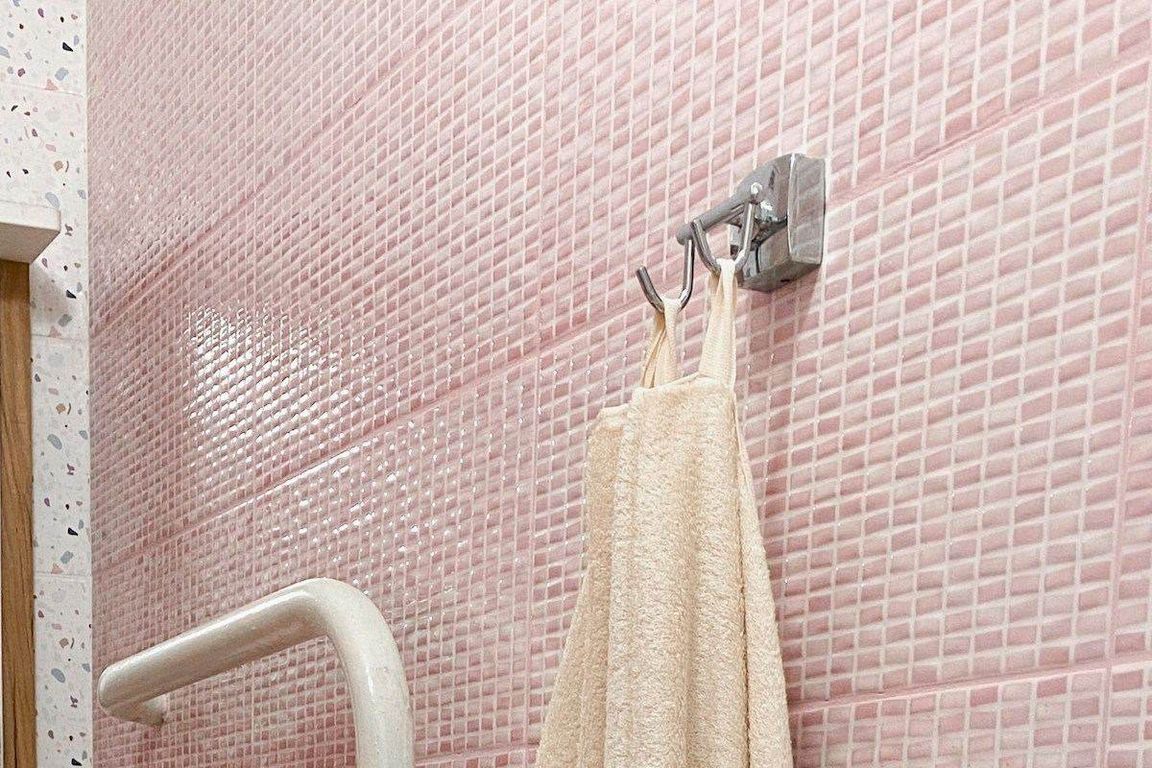 A beige bath towel hangs from a two-pronged metal hook fixed to a wall of small pink tiles, with a white towel rail at the bottom left.