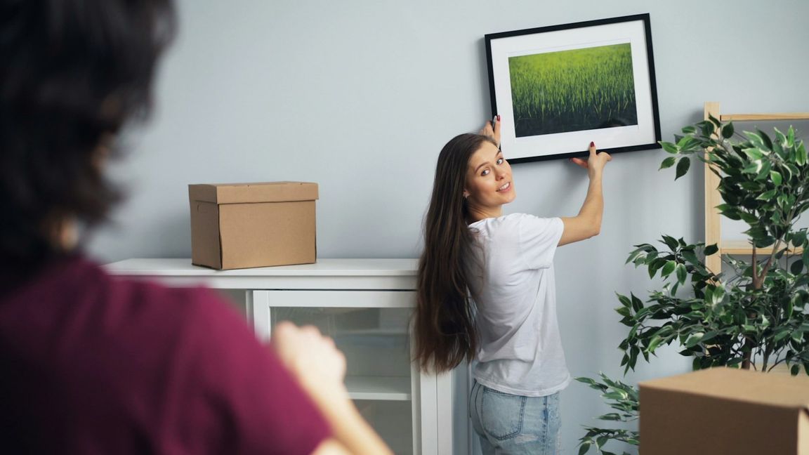A young woman in a white T-shirt hangs a black frame with a white mount on a light grey wall, surrounded by moving boxes.